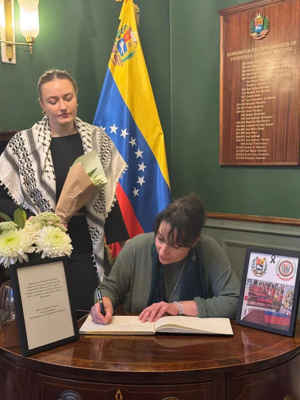 Author signing a copy of The Red New Deal for a woman holding flowers indoors near a flag and wall plaque
